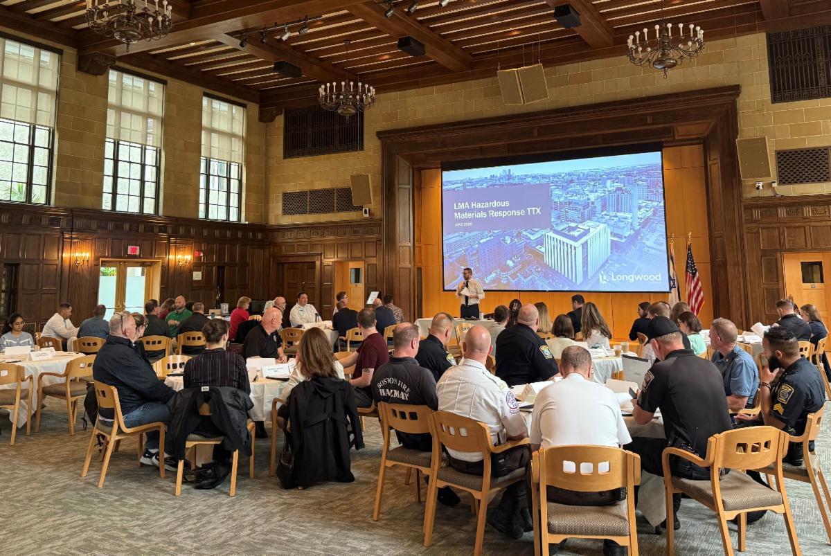 Speaker leads a room of attendees seated at tables with a PowerPoint behind him